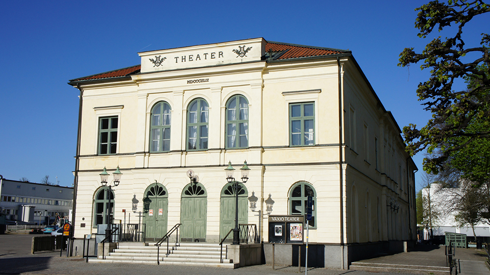 Växjö Teater. En stor vit byggnad sett snett framifrån med en blå himmel bakom.