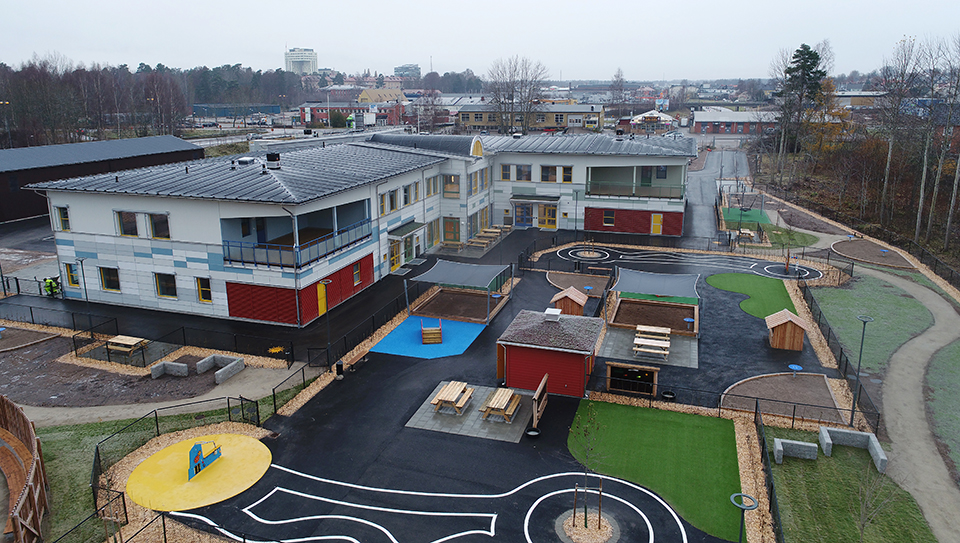 Översiktsbild av förskolan Saga. En vit byggnad med tillhörande lekplats och bebyggelse i bakgrunden (Växjö stad). Foto: Skymap.