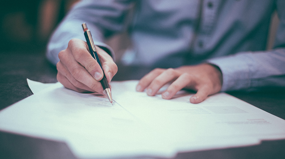 En person håller i en penna och skriver i ett dokument. Foto.