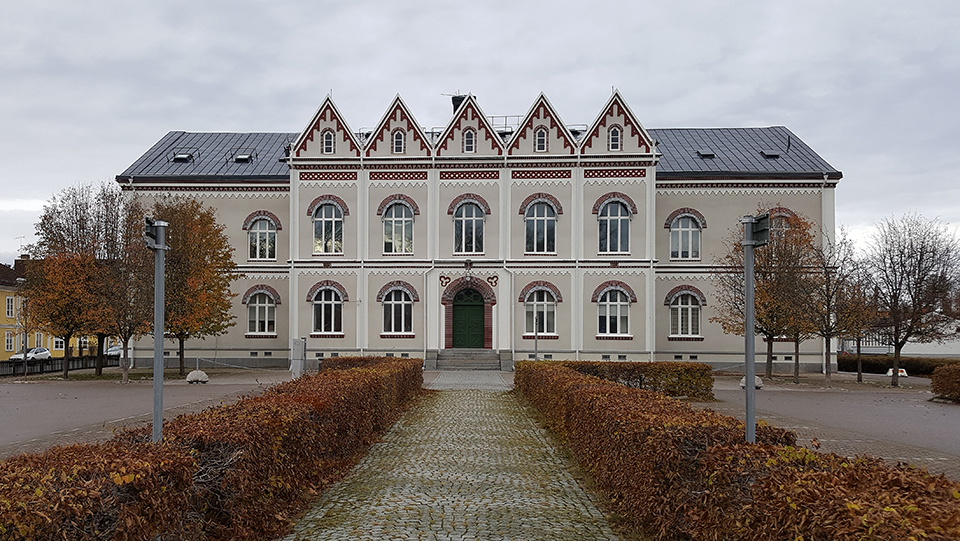Norrtullskolan, en stor grå byggnad med röda detaljer, med en gång med buskar fram till entrén. Foto.