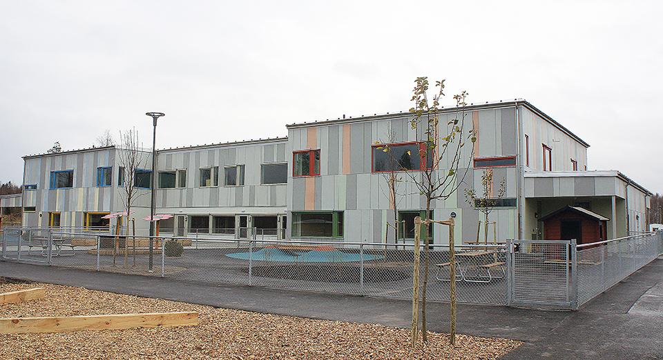 Baksidan av Vikaholms förskola med en inhägnad lekplats.Byggnaden är i två plan och till största delen grå. Foto.