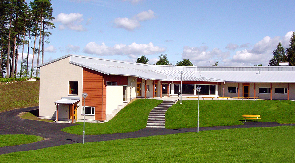 Högstorpsskolan, en byggnad i vitt och orange, med en grön gräsmatta framför och tallar i bakgrunden. Foto.